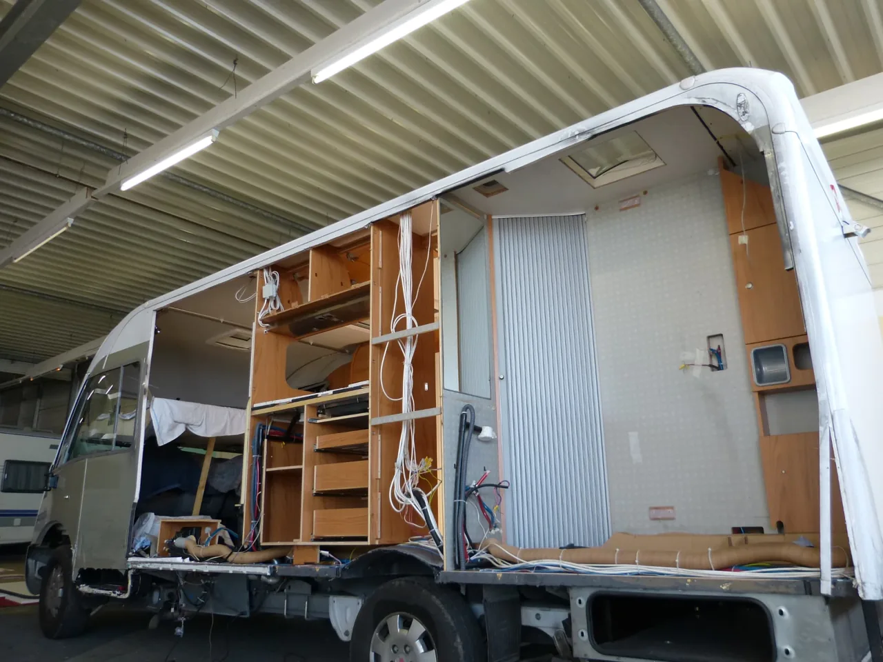Wohnmobil mit geöffneter Seitenwand, sichtbarem Innenausbau und frei liegenden Leitungen in der Werkstatthalle.