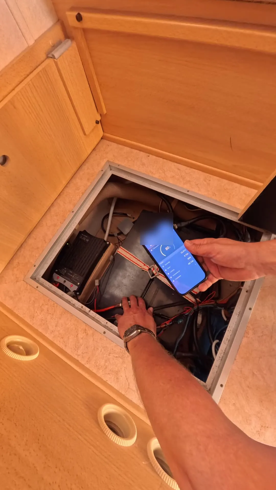 Geöffnete Bodenklappe mit elektrischen Komponenten, Leitungen und einem Smartphone in der Hand.