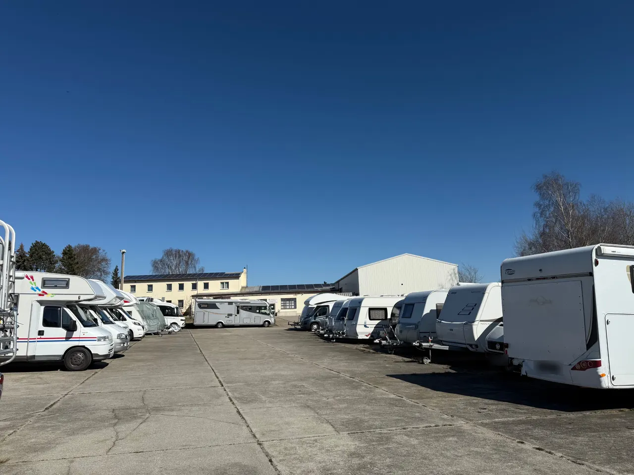Betonierte Stellfläche mit abgestellten Wohnmobilen und Wohnwagen in zwei Reihen, dahinter Betriebsgebäude.