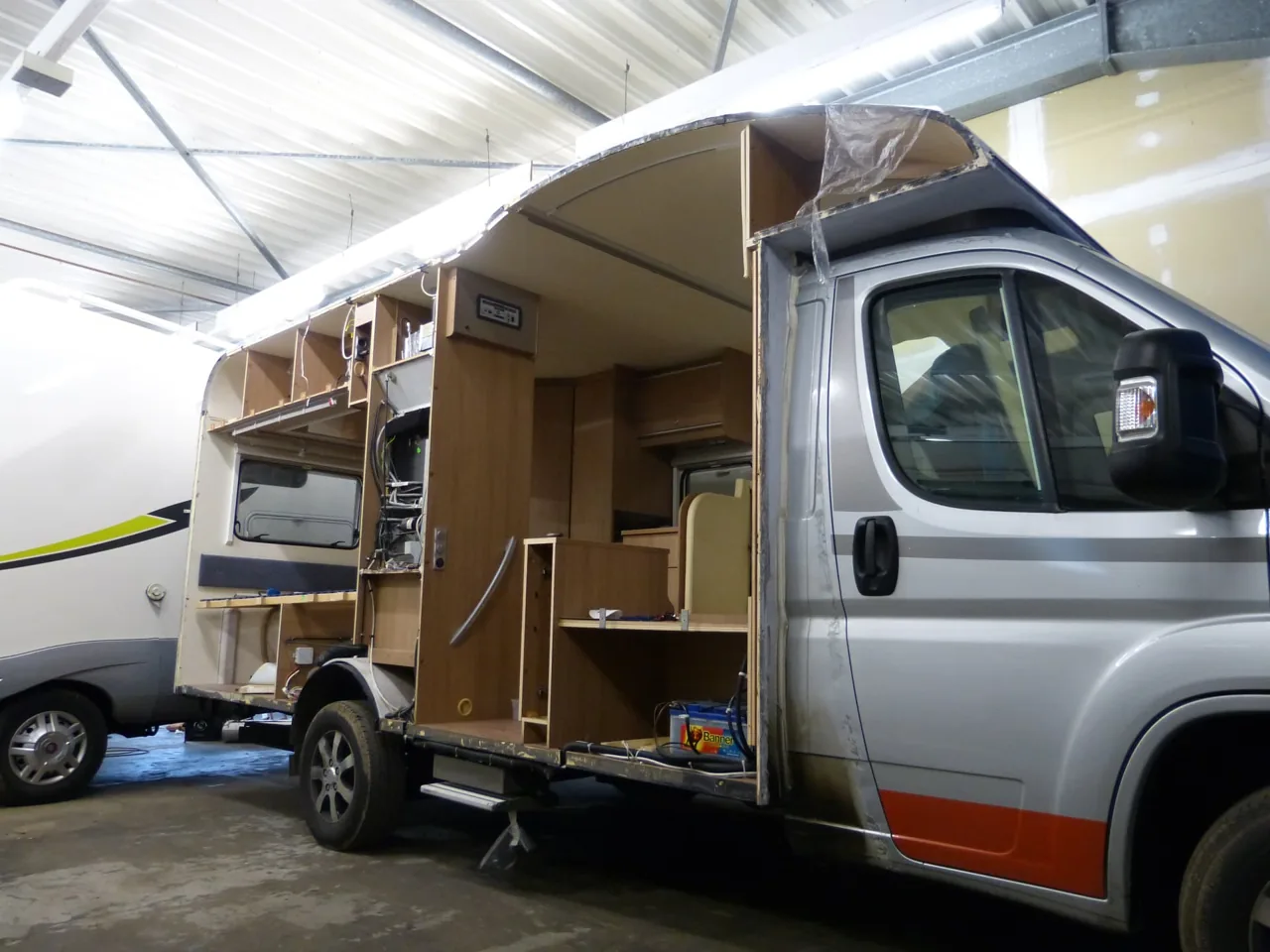 Teilweise geöffneter Wohnmobilaufbau mit sichtbaren Möbeln, Leitungen und Dachkonstruktion in der Werkstatthalle.